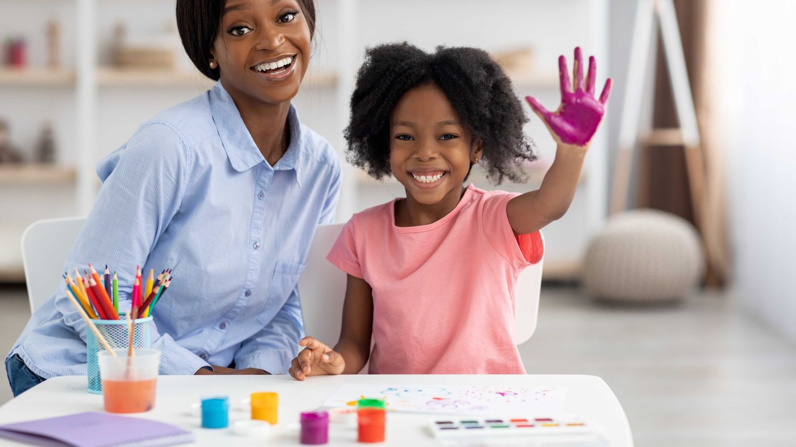 A counselor and a young girl at a craft table, the girl proudly showing her paint-covered palm