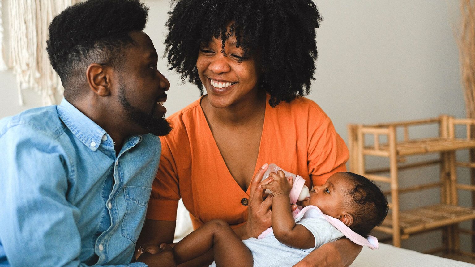 Two smiling parents sit close together on a bed, one feeding a bottle to their baby
