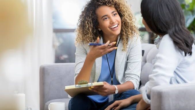 A clinician with a clipboard and lanyard talks warmly with someone seated across from her