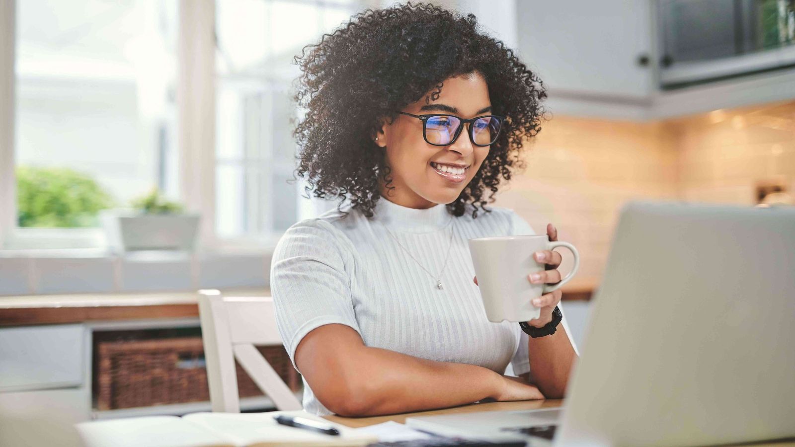 A smiling woman holding a mug at her kitchen table, talking with someone on her laptop