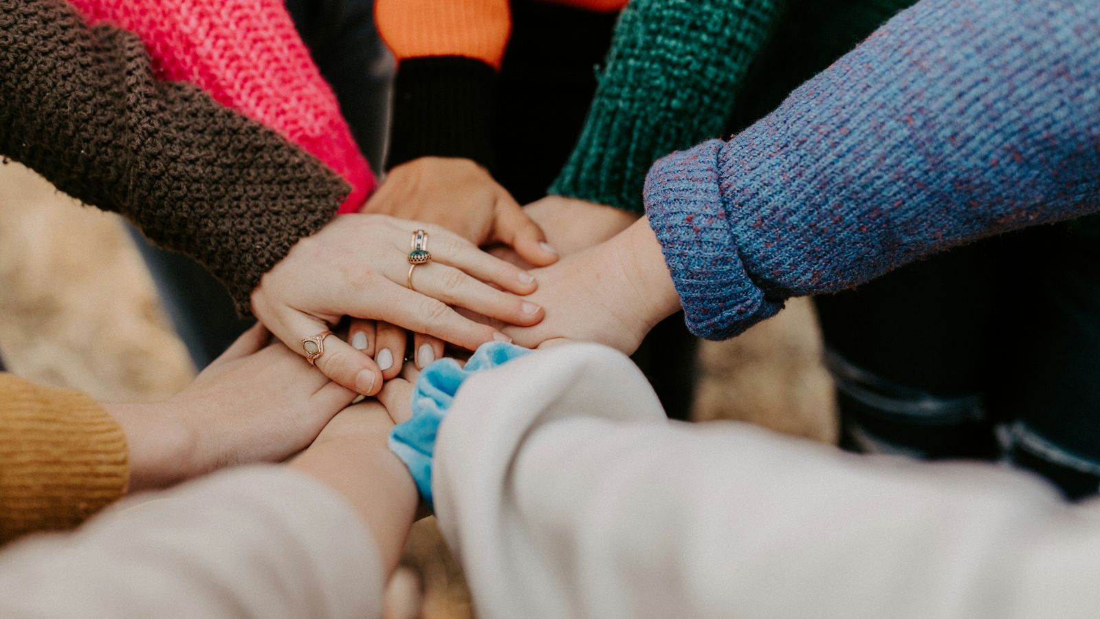 Several people in colorful knit sweaters stack their hands together in the middle of a circle