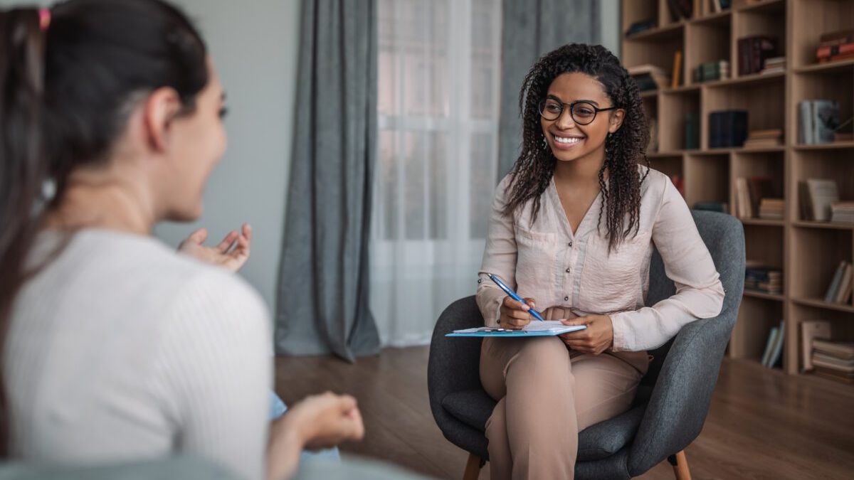 A smiling counselor with a notepad listens as a woman speaks, in a sunlit room lined with bookshelves