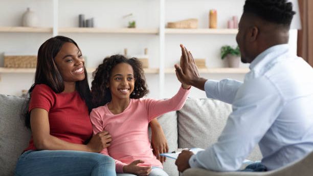 A girl high-fives a counselor across a bright office while an adult sits beside her, smiling