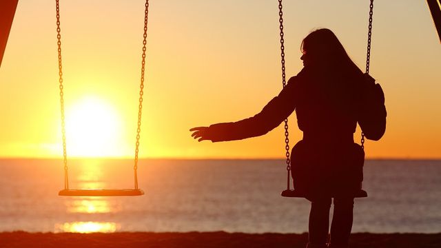 A person silhouetted on a beach swing at sunset reaches toward the empty swing beside them