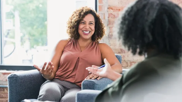 A woman talks and gestures warmly from an armchair while another person listens, pen in hand