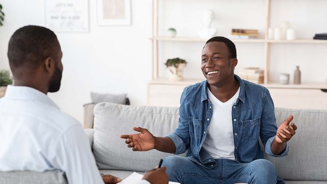 A young man on a sofa smiles and gestures mid-conversation as a counselor takes notes
