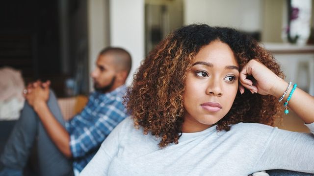 A woman sits quietly on a couch looking away while a man behind her faces the other direction