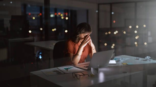 A woman works late alone at a lit desk in a dark office, hands pressed to her brow