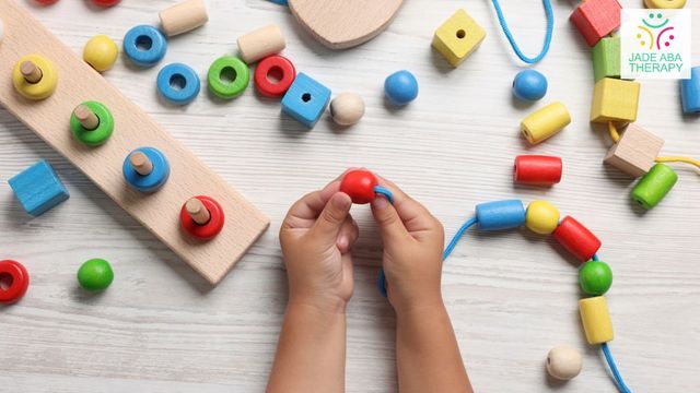 A child's hands threading a red wooden bead onto a lace, surrounded by bright stacking toys