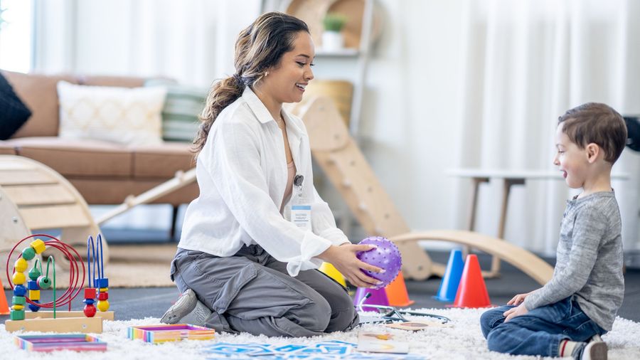 A smiling therapist kneels on a playroom rug, offering a purple ball to a laughing young boy