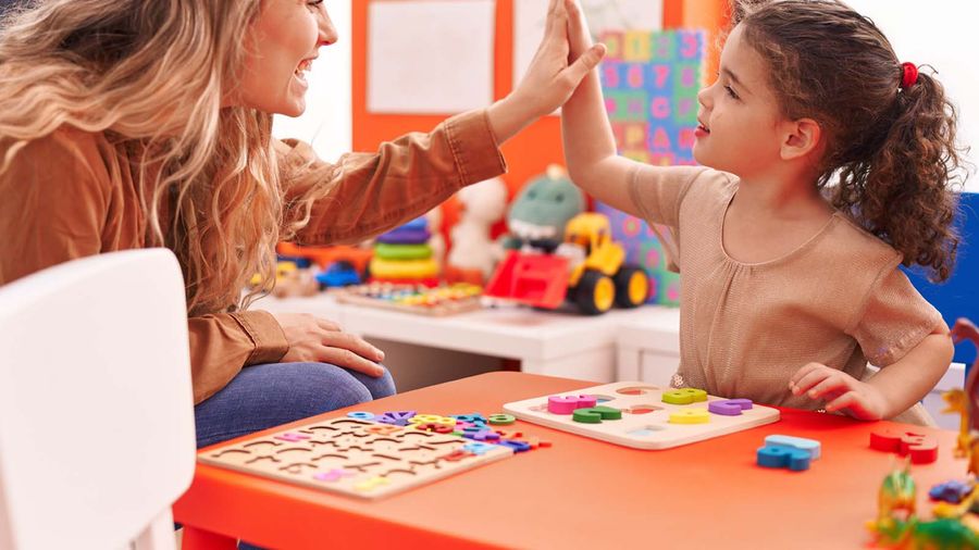 A woman and a young girl high-five over an orange table covered with letter puzzles in a playroom