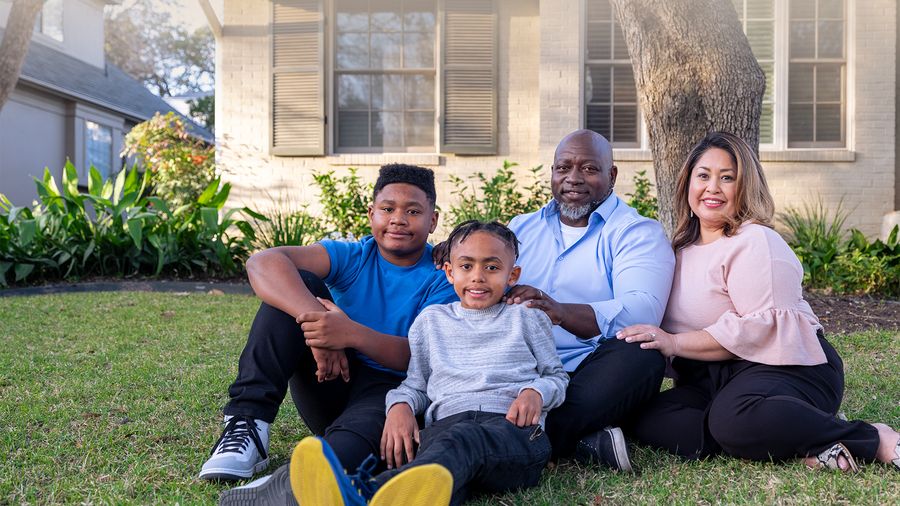 Two adults and two boys sit close together on a sunlit front lawn, smiling at the camera