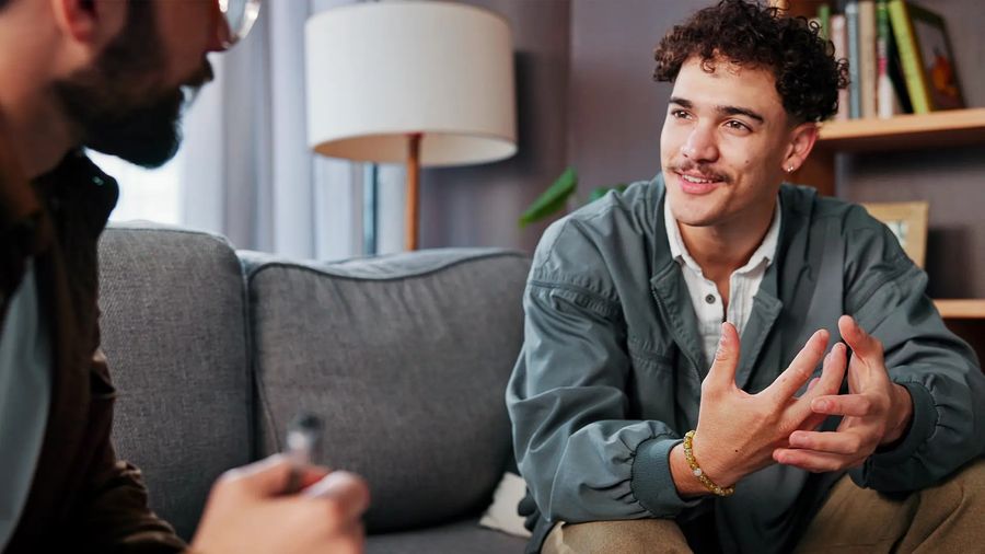 A young man gestures as he talks with a counselor holding a pen in a warm living room