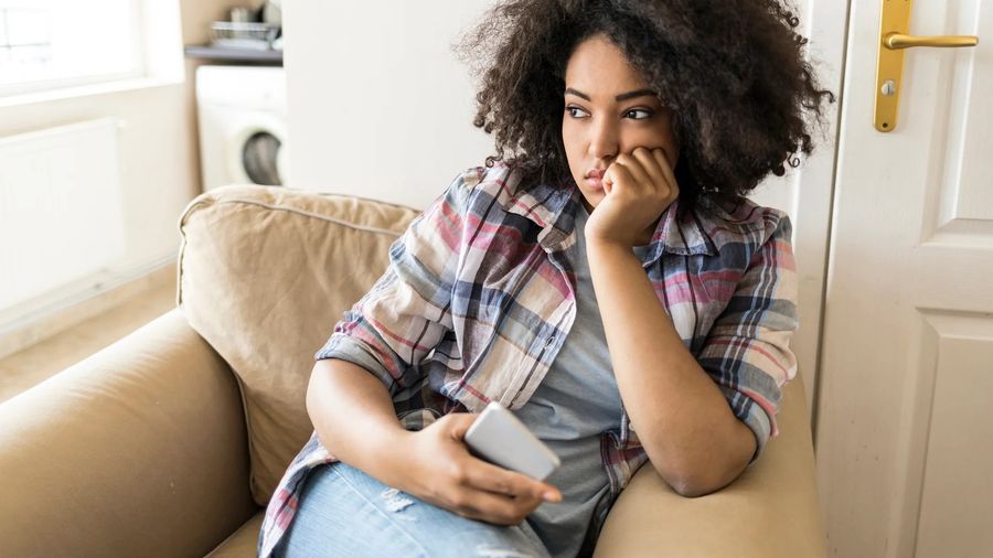 A young woman sits on a sofa with her chin on her hand, phone in hand, gazing away
