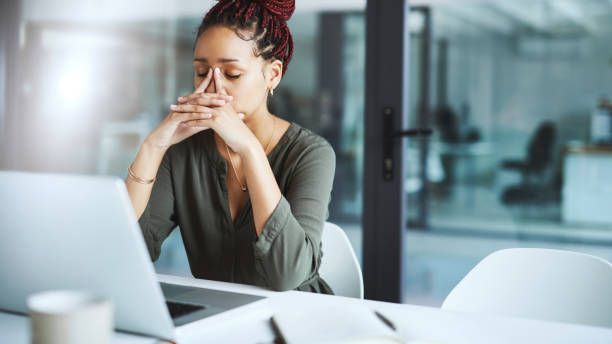 A woman at an office desk pauses with her eyes closed and hands pressed against her face
