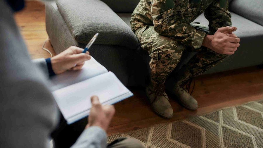 A counselor takes notes while a person in military fatigues sits on a sofa, hands clasped
