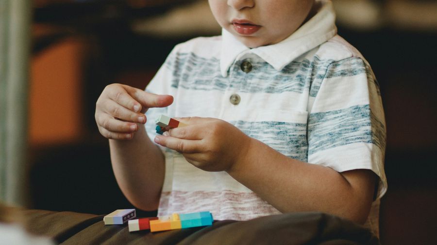 A young child in a striped polo shirt sits on a sofa, absorbed in building with coloured bricks