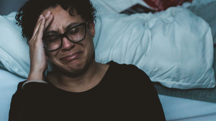 An adult in glasses sits beside a bed in dim light, one hand at the forehead, looking upset