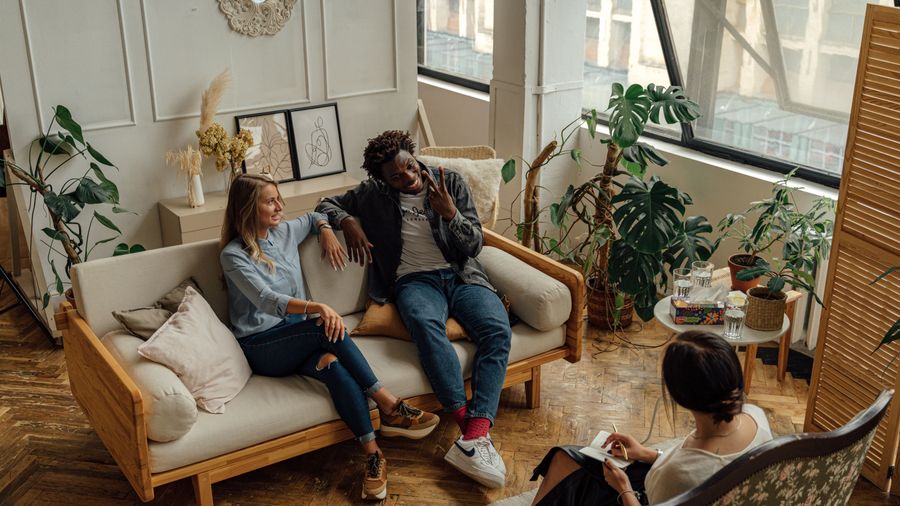 Seen from above, a couple talk and laugh on a sofa while a therapist listens and takes notes