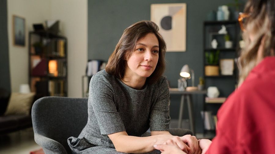 Two adults sit facing each other in a warmly lit room, one holding the other's hand reassuringly