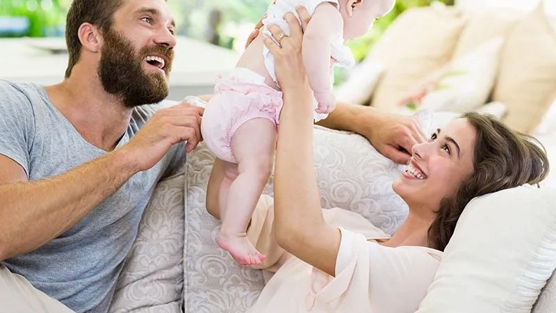 Two smiling parents lift a baby between them while relaxing on a sofa in a sunlit room