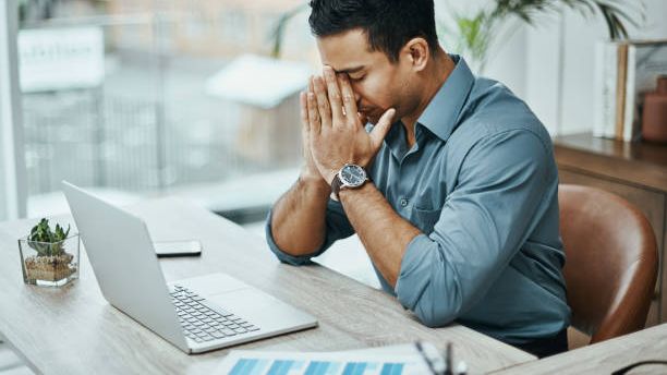 A man pauses at his desk beside an open laptop, hands pressed together against his face